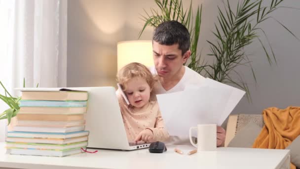 親のためのホームオフィス 遠隔で働いている父 ラップトップで作業している白いTシャツを着ている不幸な男と 自宅で彼を邪魔する幼児の赤ちゃんとテーブルに座っている文書 — ストック動画
