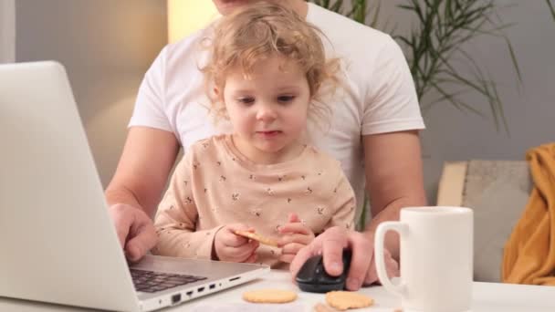 かわいいブロンドの重い髪の小さな赤ん坊の少女は 自宅でテーブルでラップトップの男に取り組んで座って 魅力的な子供は 彼のリモート仕事をしているキーボードにタイピングクッキーの父親を食べます — ストック動画