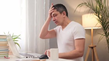 Çok çalışan beyaz tişörtlü beyaz beyaz bir adam evde dizüstü bilgisayarla çalışıyor baş ağrısı çekiyor ağrılı baş ağrısına dokunuyor çevrimiçi eğitimden yorgun düşerek dinleniyor.