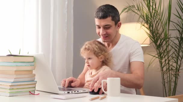 父親のゲーマーは ビデオゲームをプレイし 子供の赤ちゃんが自宅でテーブルに座っているラップトップを使用して白いTシャツを着ている子猫の娘の男と時間を過ごします — ストック動画