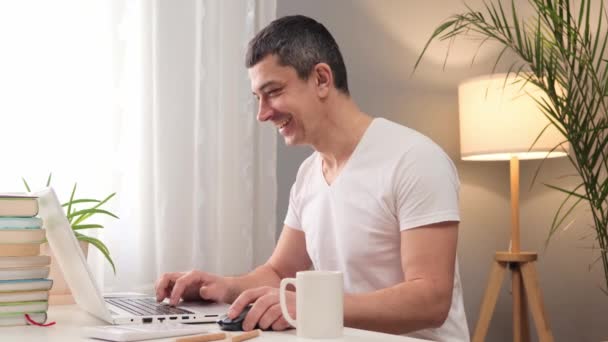 一个积极迷人的白种人 穿着白色的T恤 坐在家里的笔记本电脑上打字 做着遥远的自由职业工作 享受着他的工作 — 图库视频影像