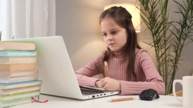 Okul çocukları için uzaktan eğitim. Küçük bir öğrenci için çevrimiçi rota. Kafkasyalı küçük kahverengi saçlı kız evde bilgisayar kullanarak bilgi yazıyor.