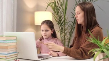 Anne çocuğa ödevlerinde yardım ediyor. Çocuk eğitiminde bir teknoloji. Ebeveyn ve çocuk çevrimiçi çalışıyor. Ödevler için dizüstü bilgisayar kullanımı. Kafkasyalı anne ve küçük kız masada dizüstü bilgisayarlarıyla oturuyorlar.