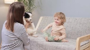 Kanepede oturan küçük, neşeli sarışın kız bebek annesi siyah benekli küçük beyaz köpeğe neşeli bir hediye ve komik tüylü bir sürpriz veriyor.