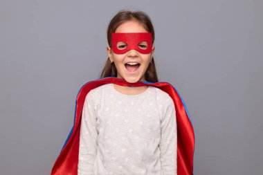 Heyecanlı, neşeli, süper kahraman kostümü giyen ve maskesini gri arka planda saklayıp mutluluk ve olumlu duygularla haykıran küçük bir kız.