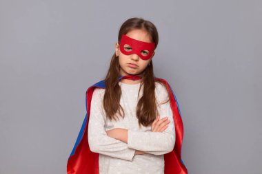 Üzgün, süper kahraman kostümü giymiş ve maske takmış üzgün küçük bir kız. Gri arka planda izole edilmiş. Kollarını kavuşturmuş, dudaklarını bükmüş üzgün ve üzgün bir şekilde dikiliyor.