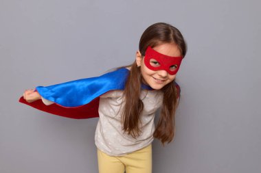 Neşeli, neşeli, süper kahraman kostümü giyen ve gri arka planda soyutlanmış maske takan küçük kız sihirli mavi ceketiyle kameraya gülümsüyor.