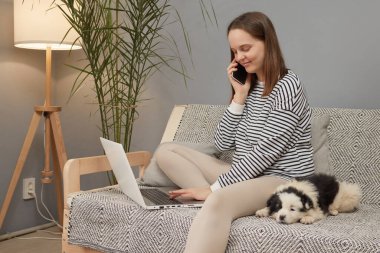 Güzel, beyaz bir kadın çizgili gömlek giyiyor. Yavru köpeğiyle koltukta oturuyor. Cep telefonuyla konuşuyor ve bilgisayarını kullanıyor.