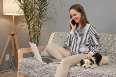 Sırıtkan, beyaz, çizgili tişörtlü bir kadın. Yavru köpeğiyle koltukta oturuyor. Bilgisayar kullanıyor ve akıllı telefondan konuşuyor.