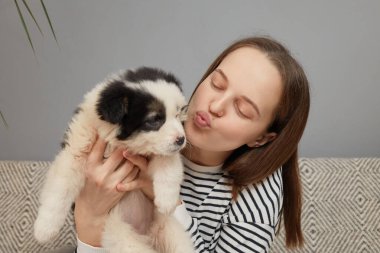 Çizgili tişört giymiş kahverengi saçlı kadın evdeki kanepede otururken küçük köpeğini kucaklarken dudaklarını büküp büyüleyici köpeğini öpüyor.