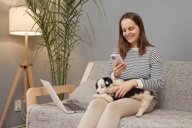 Çizgili tişört giyen beyaz bir kadın yavru köpeğiyle birlikte kanepede oturmuş internette gezinmek için dizüstü bilgisayar ve akıllı telefon kullanıyor.
