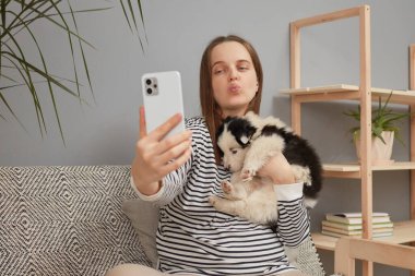 Çizgili tişört giyen, sevimli genç bir kadın. Küçük köpeğiyle blogu için akıllı telefonuyla selfie çekiyor. Kanepede oturmuş, telefon görüşmesi için hava öpücüğü yolluyor.