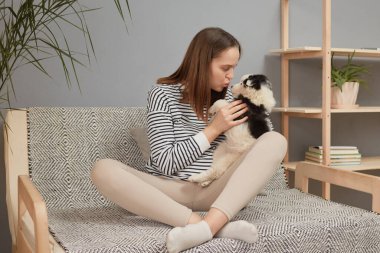 Kanepede çizgili tişört giyen komik beyaz kadın oturma odasında küçük köpeğiyle oynayıp bacak bacak bacak üstüne atarak komik hayvanını öpüyor.