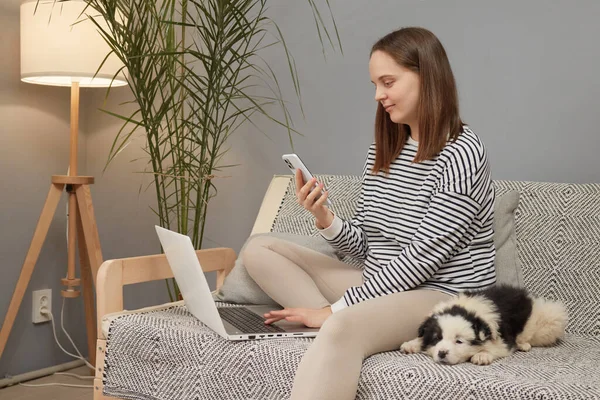 Çekici beyaz kadın çizgili tişört giyiyor yavru köpeğiyle koltukta oturuyor cep telefonu dizüstü bilgisayarı kullanarak sosyal ağda sohbet ediyor.