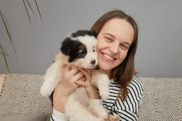 Tatmin olmuş, çizgili tişörtlü, küçük köpeğini kucaklayan sevimli genç bir kadın. Evdeki kanepede oturmuş, dişlek gülümsemesi ve mutlu ifadesiyle kameraya bakıyor.