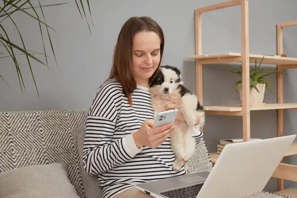 Winsome sakin, rahat, beyaz kadın çizgili tişört giymiş yavru köpeğiyle kanepeye oturmuş cep telefonuyla internette sohbet ediyor sosyal medya sayfalarını kontrol ediyor.