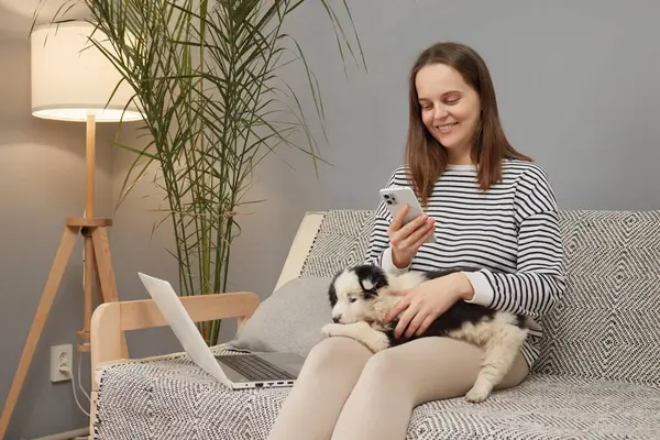 Çizgili tişört giyen beyaz bir kadın yavru köpeğiyle birlikte kanepede oturmuş internette gezinmek için dizüstü bilgisayar ve akıllı telefon kullanıyor.