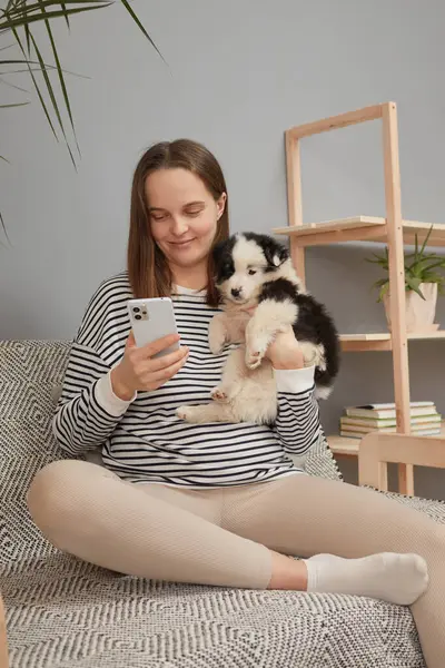 Güzel, genç, yetişkin bir kadın koltukta oturan sevimli bir köpekle internetteki kayan internet hatunları için cep telefonu kullanıyor çizgili tişört giyiyor ve akıllı telefon ekranına bakıyor.