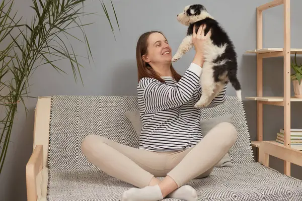 Mutlu, mutlu, beyaz, çizgili tişörtlü bir kadın kanepede oturuyor, siyah beyaz köpeğini büyütüyor ve oturma odasında poz verip eğleniyor.