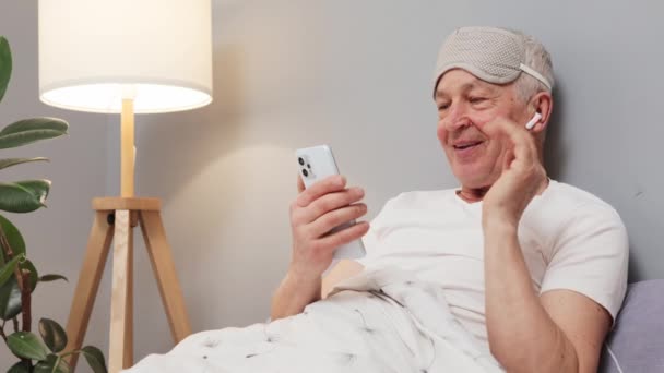 笑容满面的白种人老人 戴着睡眠面具 手里拿着摄像机在床上挥手打招呼 早上的网络通信有虚拟会议 — 图库视频影像