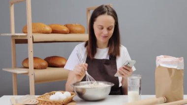 Kahverengi önlük giymiş, iş yerinde oturmuş akıllı telefon kullanarak ekmek pişirmek için malzemeleri karıştıran hoş bir kadın fırıncı.