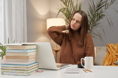 İşyerinde çalışırken kahverengi kazak giyen yorgun genç bir kadın iş yerinde rahatsız bir vaziyette otururken boynunda ya da sırtında ağrı hissediyor.