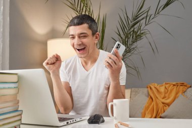 Aşırı mutlu, mutlu, beyaz bir adam dizüstü bilgisayarda çalışıyor ve ev içinde telefon kullanıyor. Başarısını kutluyor. İş yerinde yumruklarını sıkmış ve seviniyor.