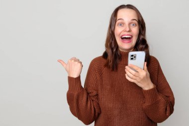 Kahverengi kazaklı, neşeli Kafkas saçlı kadın akıllı telefonla konuşuyor açık gri arka planda tek başına duran reklam alanını işaret ediyor.