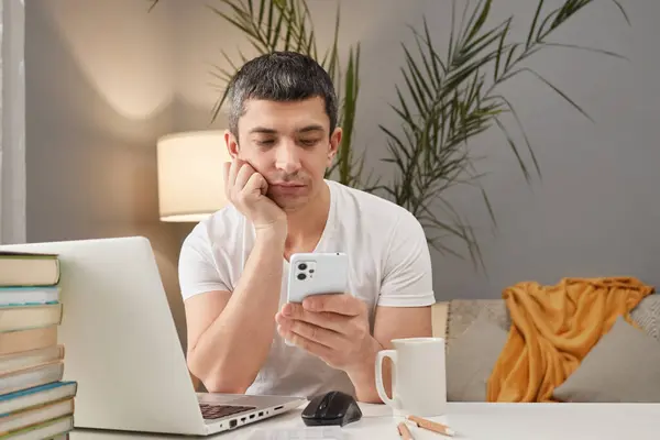 Online communication with a smartphone. Pensive thoughtful Caucasian man working on laptop and using phone in home interior freelancer taking break scrolling online while having free minutes