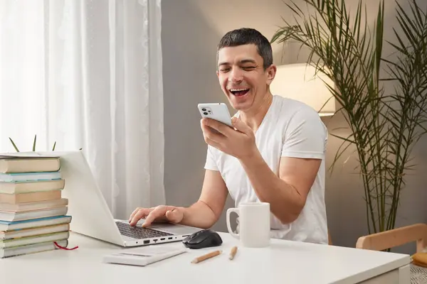 Connecting devices for efficient work. Cheerful smiling positive Caucasian man working on laptop and using phone in home interior looking at screen and laughing uses voice assistant