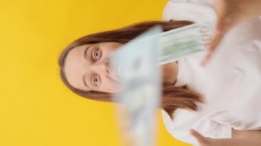Dikey video. Aşırı sevinçli beyaz tişört giyen, sarı arka planda izole edilmiş, yüksek maaş alan, banknotlar atan beyaz kadın şaşırdı.