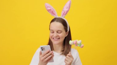 Güzel kahverengi saçlı, tavşan kulaklı bir kadın sarı arka planda izole edilmiş bir şekilde poz veriyor. Elinde akıllı telefon, internette arkadaşlarıyla sohbet ediyor ve mesajları okuyor.
