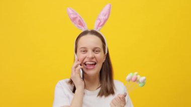 Tavşan kulaklığı takan heyecanlı beyaz kadın sarı arka planda izole bir şekilde poz veriyor cep telefonuyla konuşuyor mutlu bir şekilde gülüyor ve arkadaşlarını selamlıyor.