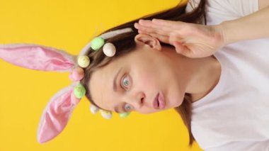 Dikey video. Tavşan kulaklığı takan meraklı genç bir kadın ve sarı arka planda izole edilmiş bir şekilde duran kek çelengi birinin fısıldadığını duymaya çalışıyor.