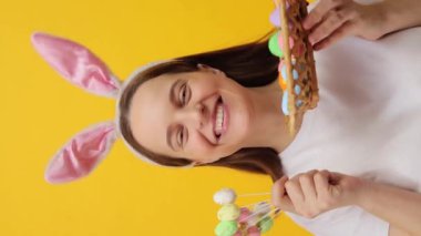 Dikey video. Tavşan kulaklığı takan güzel, mutlu, genç bir kadın. Sepette Paskalya yumurtası ve pasta kutlaması gösteriyor. Sarı arka planda izole bir şekilde tatil kutlaması yapıyor.
