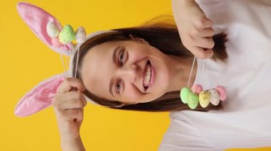 Dikey video. Tatil boyunca neşeli şenlikler. Mutlu gülümseyen yetişkin kadın tavşan kulaklığı takıyor sarı arka planda yalnız poz veriyor.