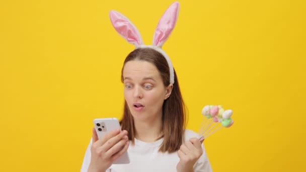 衝撃を受けた不幸な茶色の毛髪の女性は 大きな目読書ニュースで携帯電話画面を見ている黄色の背景の上に隔離されたバニーの耳のヘッドバンドを着用 — ストック動画