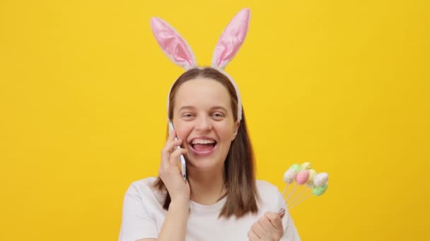 エキサイティングな白人女性ウサギの耳のヘッドバンドを着用し 携帯電話で話す黄色の背景の上に隔離され 喜んで肯定的な感情を表現する友人 — ストック動画