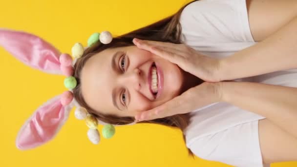 バーティカルビデオ 笑顔の白人女性ウサギの耳のヘッドバンドとケーキのポップは お祝いの気分にいる幸せな顔でカメラを見ている黄色の背景の上に隔離されたポーズ — ストック動画