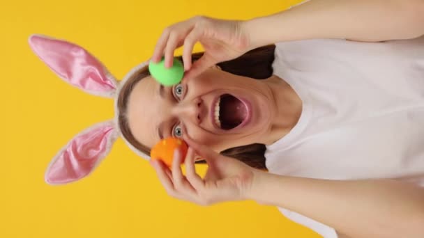 バーティカルビデオ 驚いた 興奮した若い大人の女性 バニー耳のヘッドバンドを着て 黄色の背景に隔離された 色の卵で目を覆う うわー — ストック動画