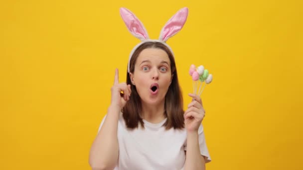 有灵感的棕色头发的女人 戴着兔子耳朵的头带 手里拿着复活节彩蛋形状的蛋糕 她非常想把节日的庆祝活动与黄色背景隔离起来 — 图库视频影像