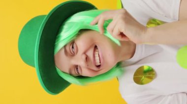 Dikey video. Parlak saçlı, yeşil cüce şapkalı, sarı arka planda izole edilmiş neşeli bir kadın St. Patrick kutlamasında mutlu bir şekilde gülümsüyor.
