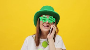 Sarı arka planda izole edilmiş yeşil şapkalı ve yonca gözlüklü neşeli St. Patrick Günü Leprikon kadın cep telefonundan arkadaşlarıyla mutlu bir şekilde gülüyor.
