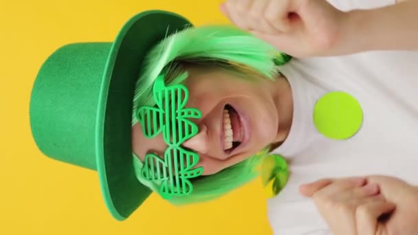 バーティカルビデオ お祝いのパーティーで幸せに踊る黄色の背景の上に隔離された緑の帽子とクローバーグラスで楽しむセントパトリックデイのレプリカンモデルに興奮 — ストック動画