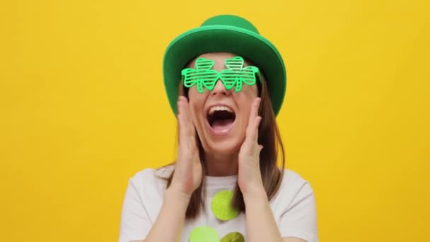 幸せな喜びの聖パトリックの日 緑の帽子と黄色の背景に隔離されたクローバーガラスのモデル女性は アナウンスを叫ぶ口の近くに手を保ちます — ストック動画
