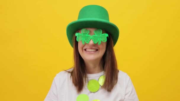 喜ばしい友好的な女性は緑色の帽子を身に着け 幸せを表現する黄色い背景の上に隔離されたクローバー メガネ はい同意します — ストック動画