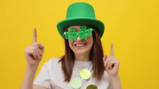 美しいポジティブなセントパトリックデイレプチャンモデル 緑の帽子とクローバーグラスで女性は インデックス指を上向きに示す黄色の背景ダンスの上に隔離 — ストック動画
