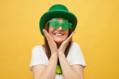 İrlanda şapkası ve yonca kutlaması. İrlanda 'da kültürel gelenekler. Yeşil leprikon şapka ve yonca gözlük takan sevimli beyaz kadın sarı arka planda tek başına duruyor.