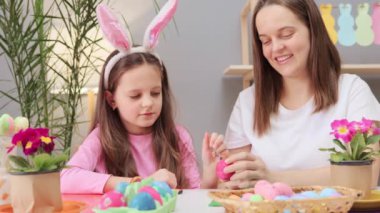 Paskalya hazırlama aktivitesi. Kahverengi saçlı küçük bir kız ve annesi bayram kutlamaları sırasında neşeli iç mekanlarda Paskalya yumurtası boyuyorlar.