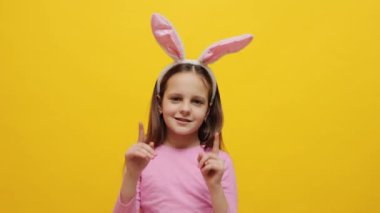 Sarı arka planda duran pembe tavşan kulaklığı takan mutlu küçük bir kız reklam için iki parmağını da yukarı kaldırıyor.